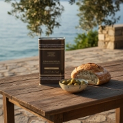 A 1000ml tin of Navarino Icons Extra Virgin Olive Oil on a rustic wooden table with fresh bread and olives, overlooking the Greek coast at sunset.