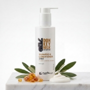 Mastic Spa 2 in 1 Shampoo and Conditioner bottle displayed on a white marble surface with fresh olive leaves, amber mastic resin tears, and a dollop of the white cream product.