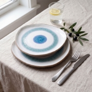 Two stacked Aegean Eye stoneware plates on a linen tablecloth with stainless steel cutlery and an olive branch.
